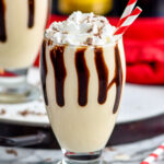 two glasses of Frozen Mudslide with chocolate drizzle, whipped cream, two straws and bottles of Kahlua and Baileys in the background