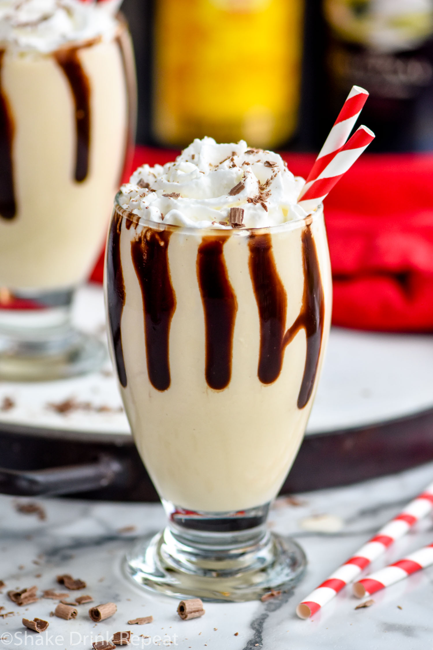 two glasses of Frozen Mudslide with chocolate drizzle, whipped cream, two straws and bottles of Kahlua and Baileys in the background