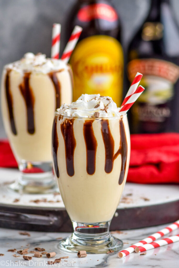 two glasses of Frozen Mudslide with whipped cream, chocolate drizzles, straws and bottles of Kahlua and Baileys in the background