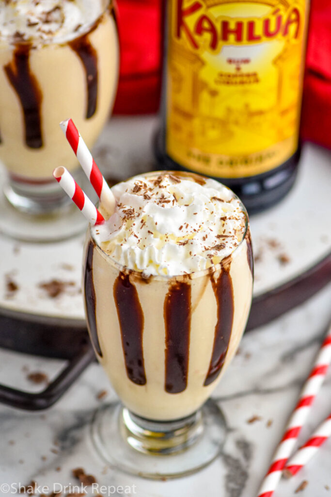 two glasses of Frozen Mudslide recipe topped with whipped cream, chocolate shavings and two straws with bottle of Kahlua in the background