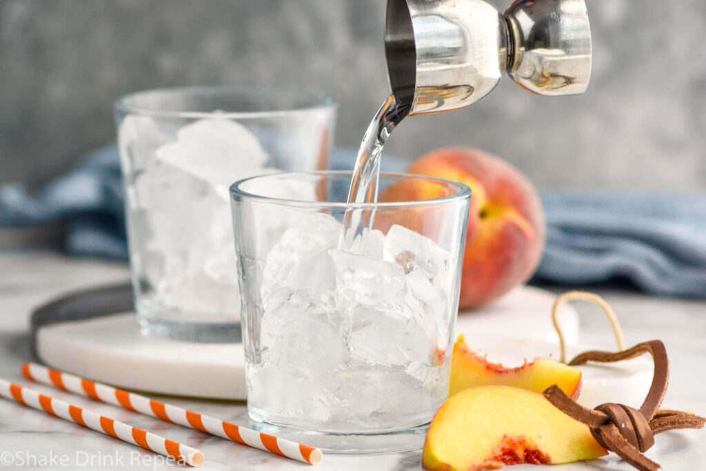 jigger of peach schnapps pouring into a glass of ice to make a Fuzzy Navel cocktail surrounded by fresh peaches and straws