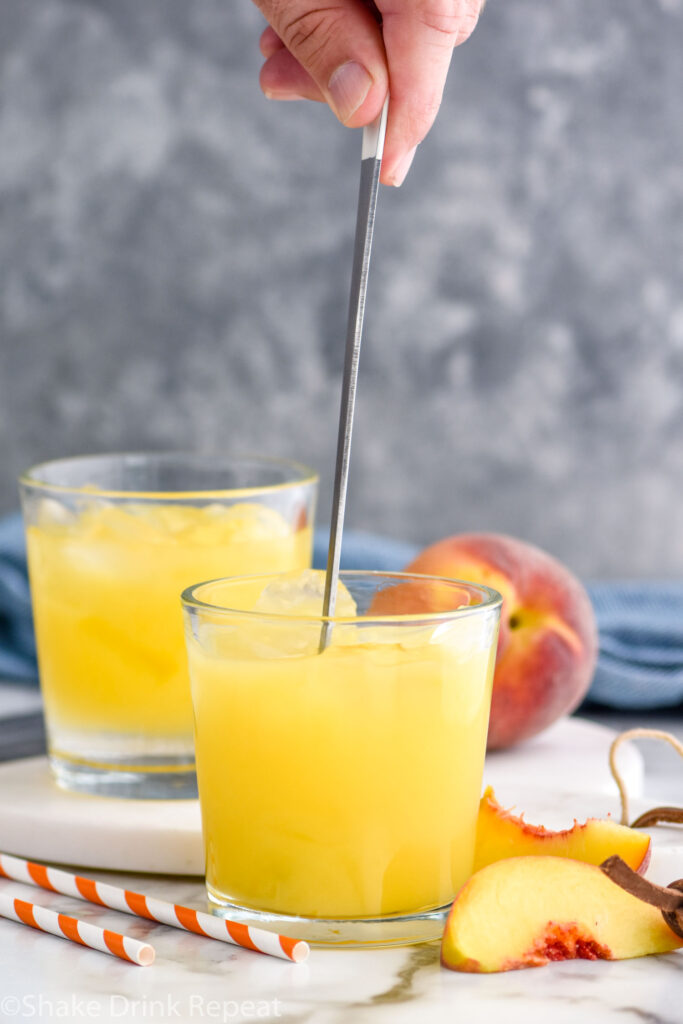 man's hand stirring glass of Fuzzy Navel with ice surrounded by fresh peach slices and straws