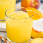 two glasses of Fuzzy Navel with ice and orange slice garnish surrounded by fresh peach slices and a straw