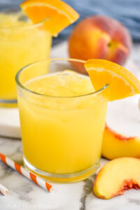 two glasses of Fuzzy Navel with ice and orange slice garnish surrounded by fresh peach slices and a straw