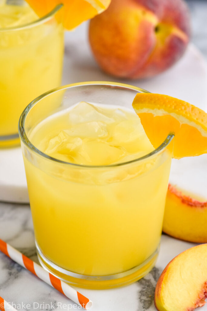 two glasses of Fuzzy Navel cocktail with ice and orange slice garnish surrounded by fresh peach slices and a straw