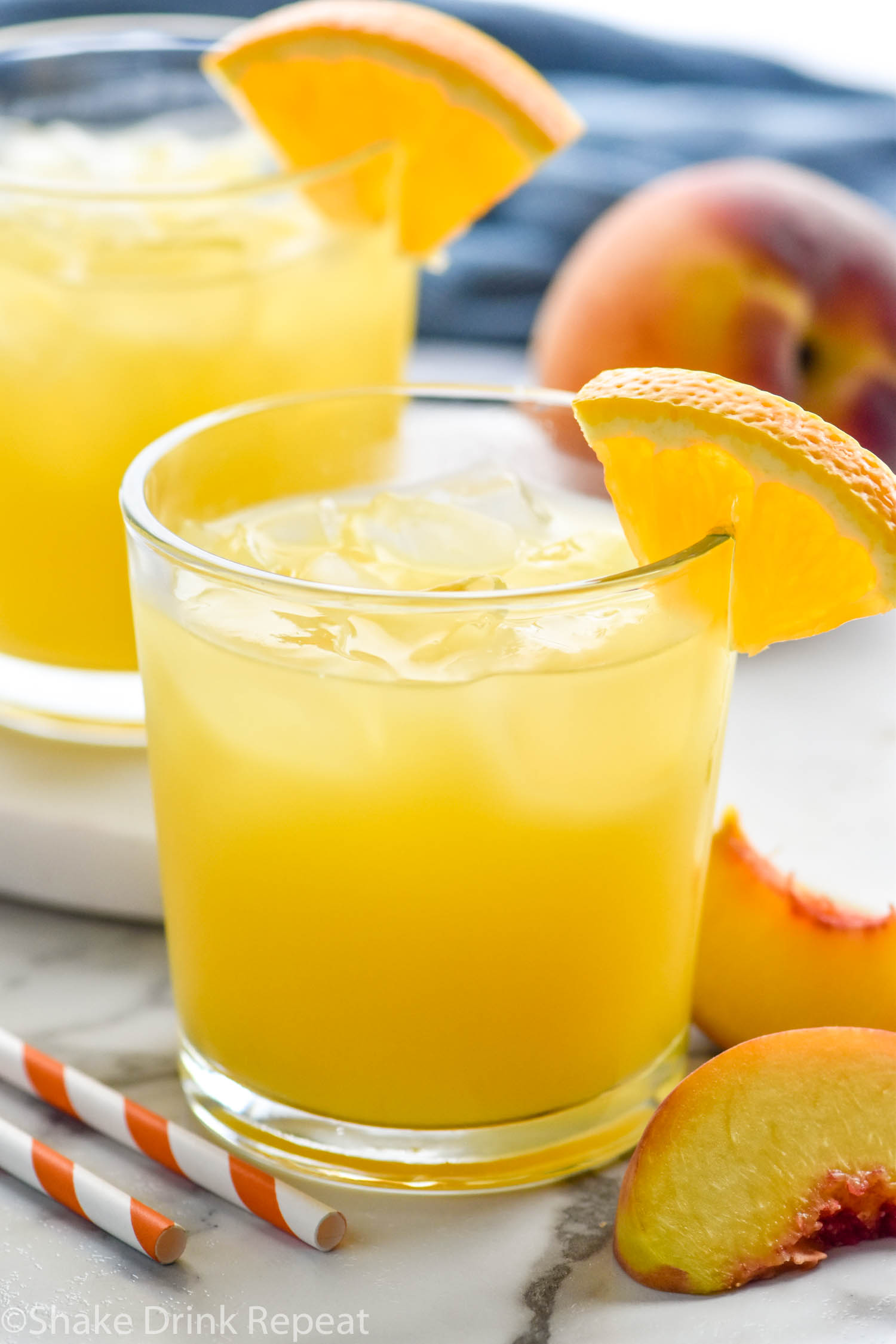 two glasses of Fuzzy Navel with ice and orange slice garnish surrounded by fresh peaches and two straws