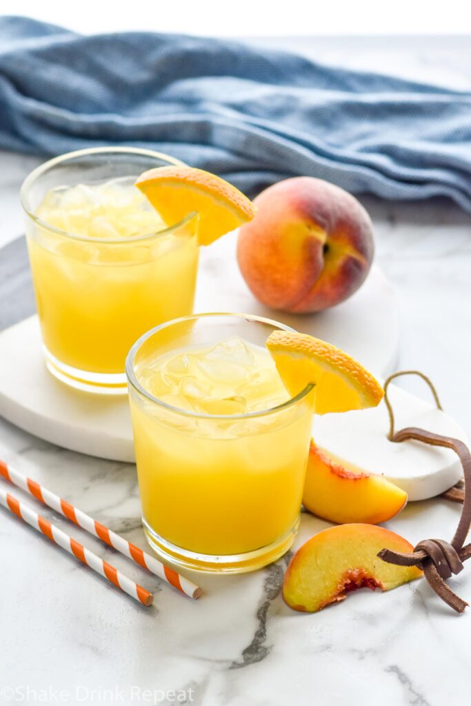 two glasses of Fuzzy Navel with ice and orange slice garnish surrounded by fresh peaches and two straws