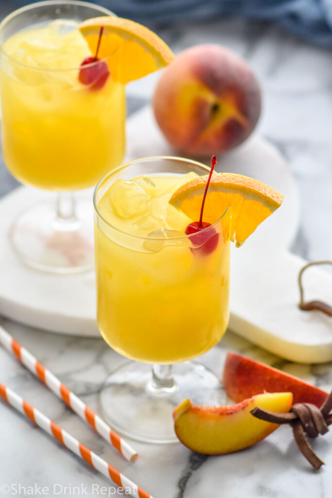 two glasses of Hairy Navel with ice, garnished with a cherry and slice of orange, and surrounded by straws and fresh peaches