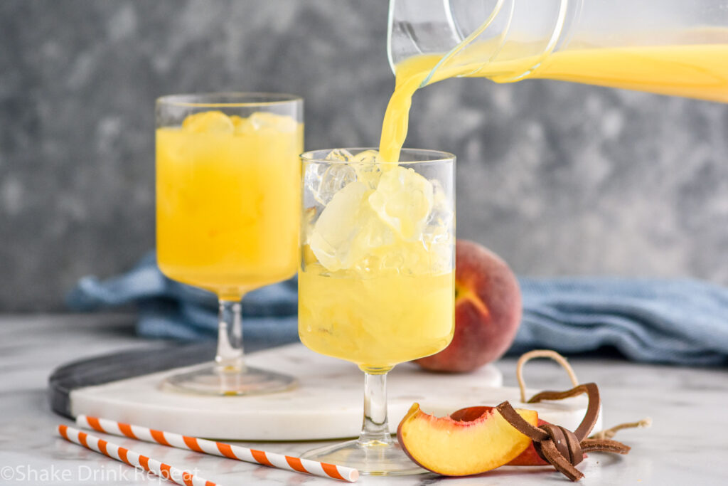 pitcher of orange juice pouring into a glass of Hairy Navel recipe with ice surrounded by fresh peaches and two straws
