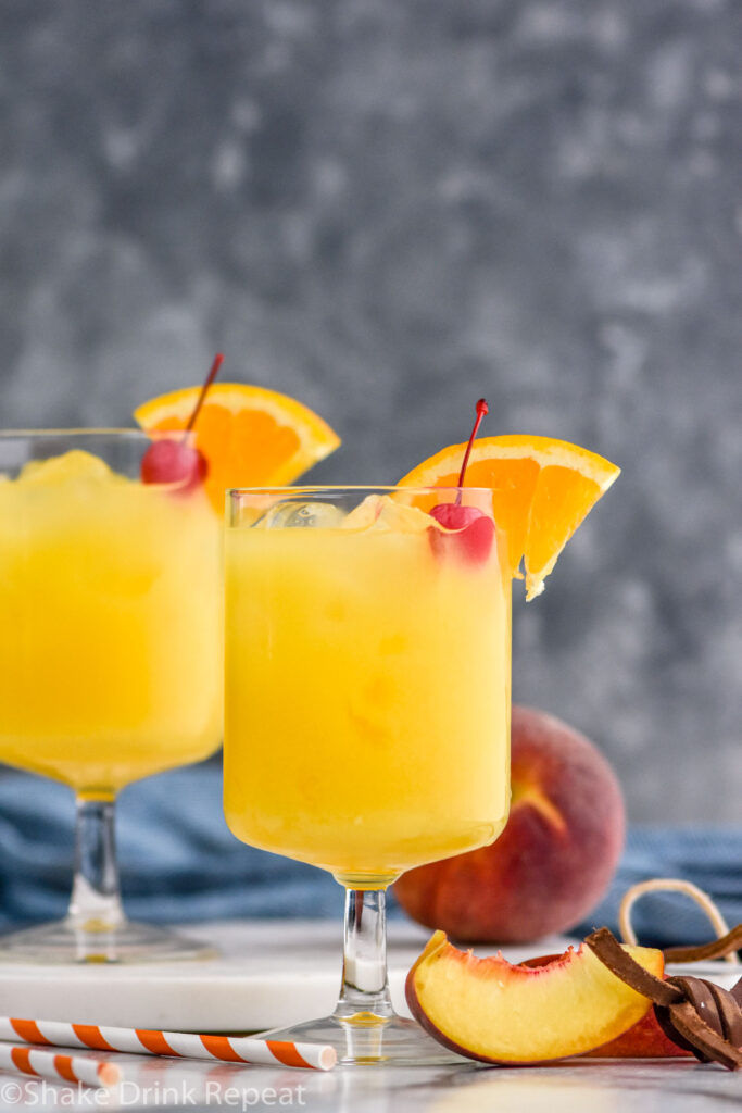 two glasses of Hairy Navel with ice, garnished with a cherry and orange slice, surrounded by straws and fresh peach