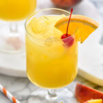two glasses of Hairy Navel cocktail with ice and garnished with a cherry and slice of orange, surrounded by a straw and fresh peaches