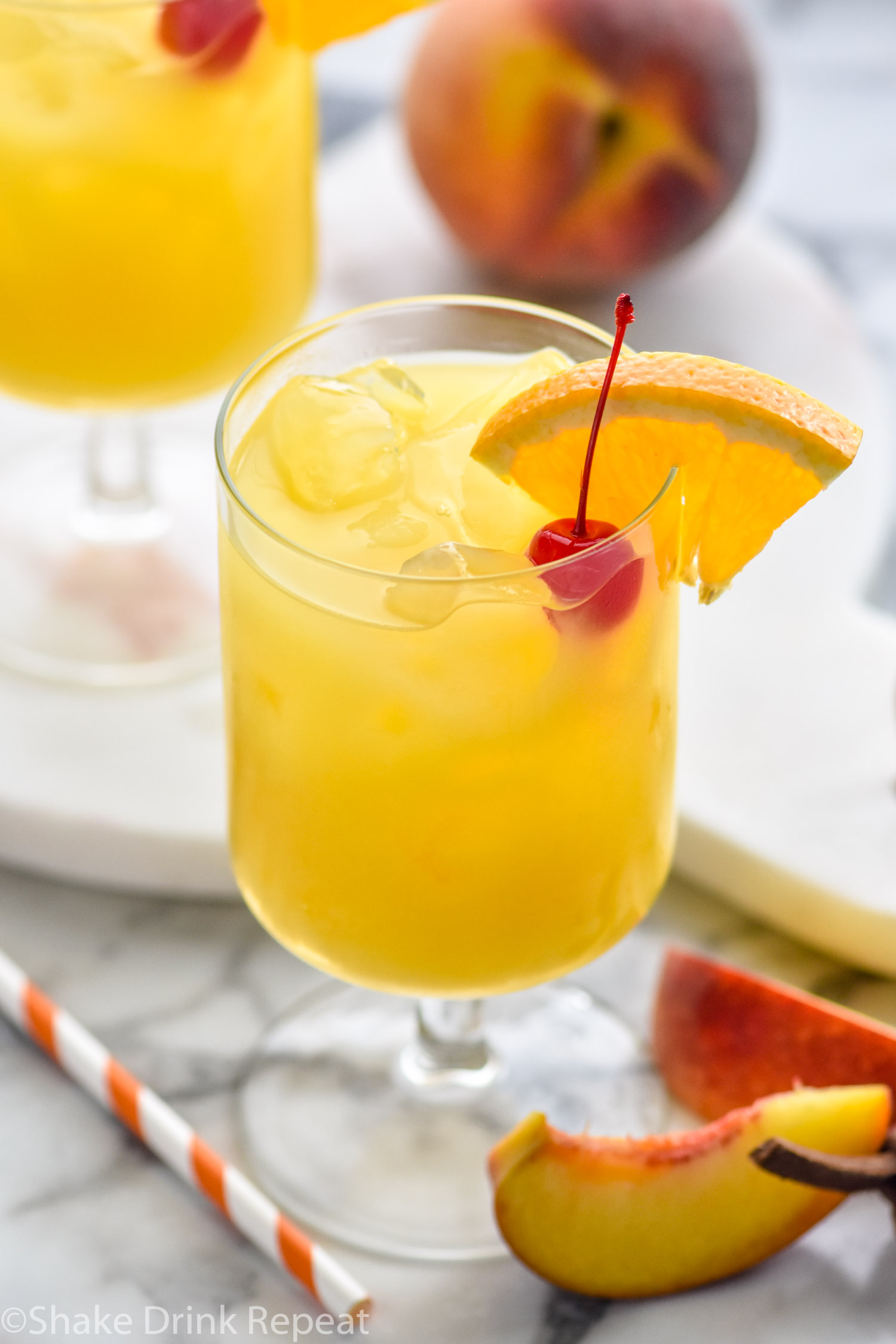 two glasses of Hairy Navel cocktail with ice and garnished with a cherry and slice of orange, surrounded by a straw and fresh peaches