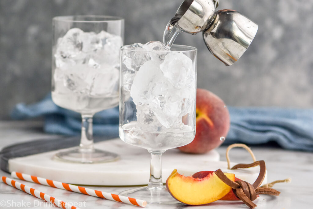 two glasses of ice with jigger of vodka pouring into glass to make a Hairy Navel recipe surrounded by straws and fresh peaches