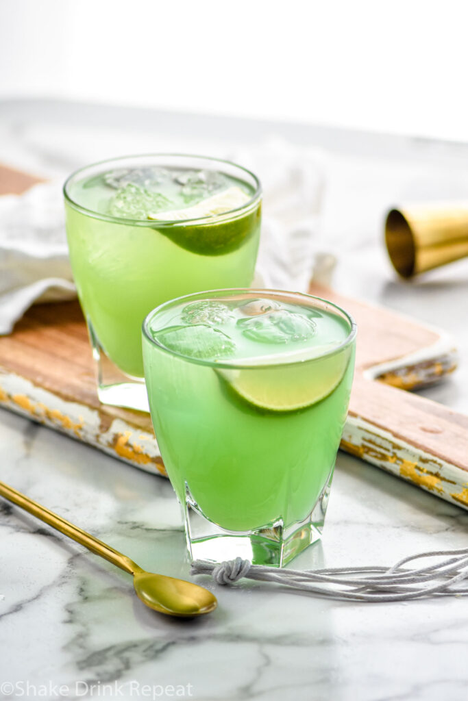 two glasses of incredible hulk cocktail with ice and lime wedge garnish, surrounded by gold spoon and jigger