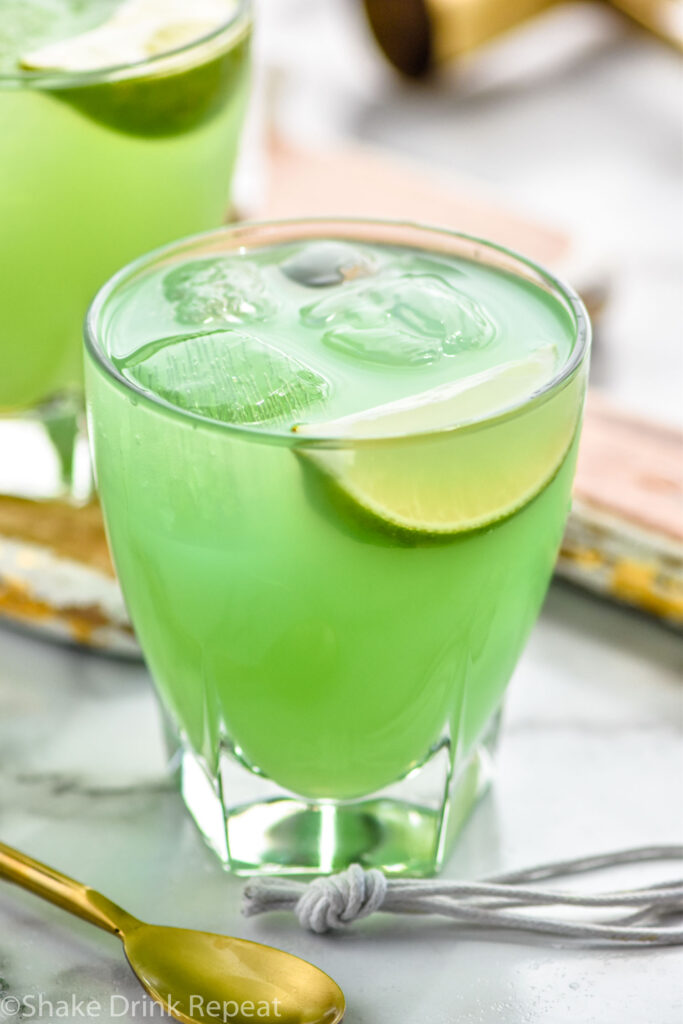 two glasses of incredible hulk cocktail with ice and lime wedge with a spoon laying in front