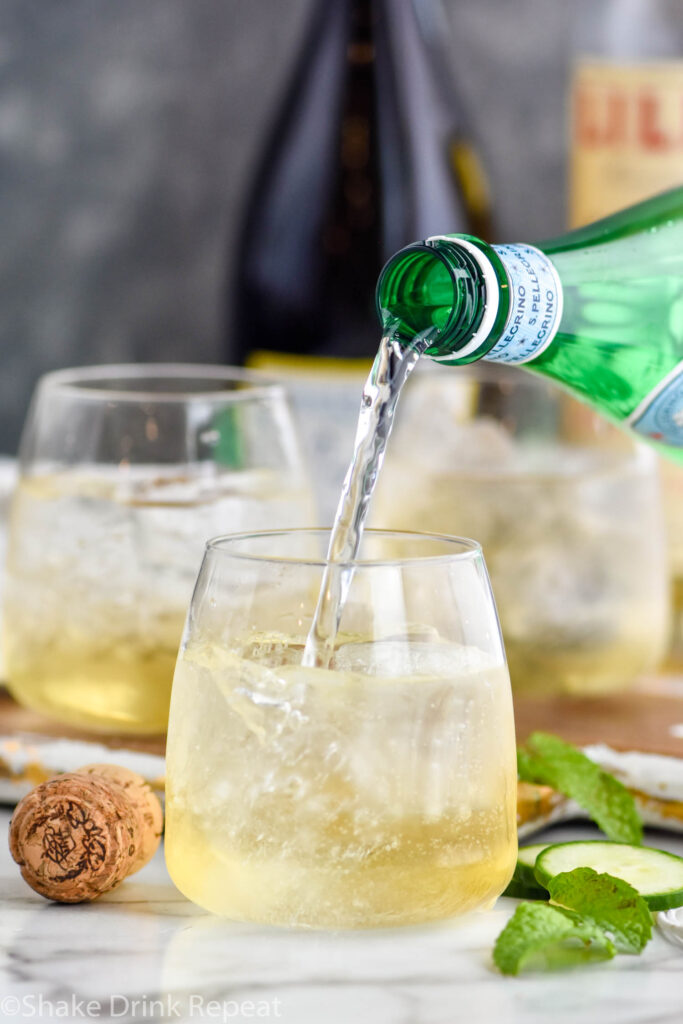 bottle of club soda pouring into a glass of Lillet Spritz with ice, fresh mint leaves, and cucumber slices. Bottle of Prosecco and Lillet Blanc in the background