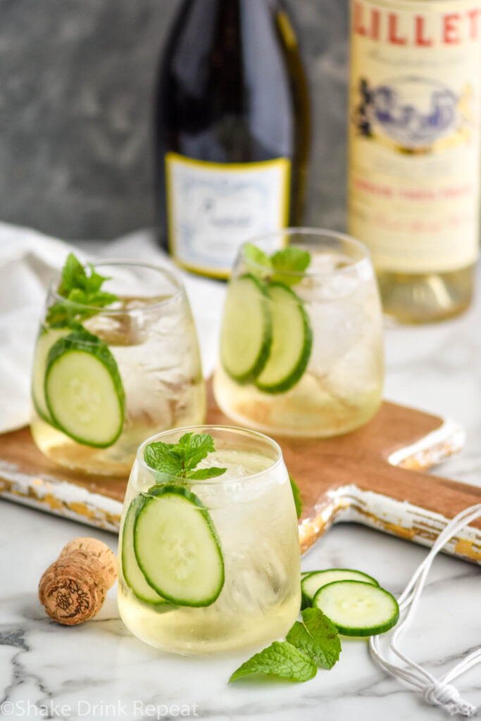 three glasses of Lillet Spritz with ice, fresh mint leaves, and cucumber slices with bottles of Prosecco and Lillet Blanc in the background