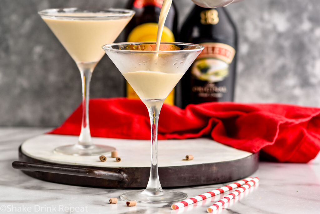 pouring Mudslide recipe into a martini glass surrounded by chocolate shavings, two straws, a bottle of Bailey's, and a bottle of Kahlua