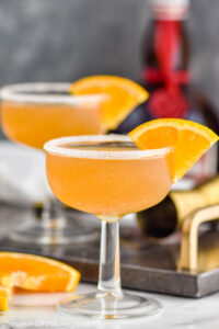 two glasses of Sidecar cocktail with sugared rim, fresh orange slice garnish and bottle of Cognac in the background