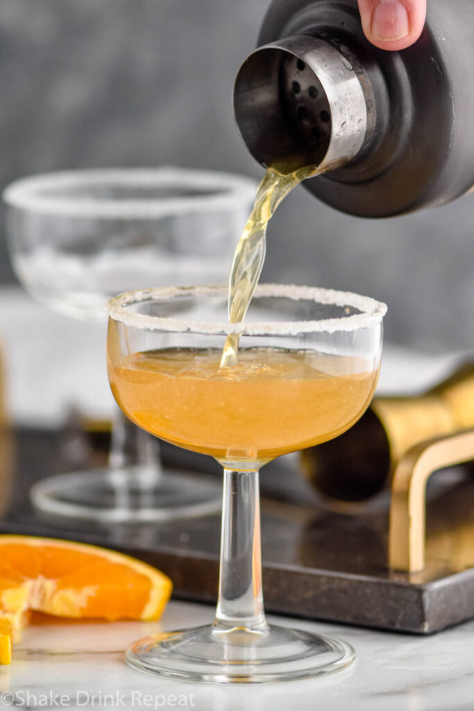 shaker of Sidecar ingredients pouring into a glass with sugared rim surrounded by orange slice