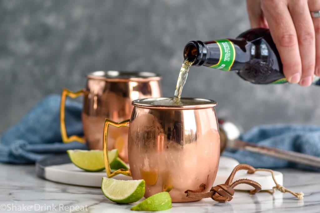 man pouring bottle of ginger beer into a copper mug of Tequila Mule ingredients surrounded by fresh lime wedges