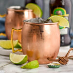 two copper mugs of tequila mules with ice, fresh lime wedges, and bottles of ginger beer