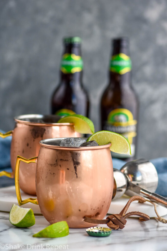 two copper mugs of tequila mules with ice, lime, and two bottles of ginger beer in the background