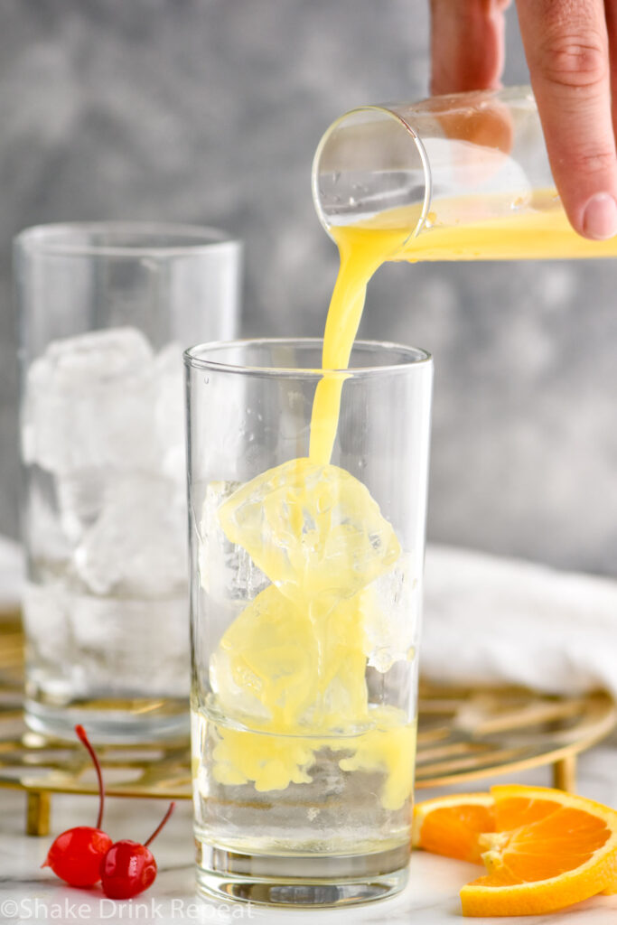 man pouring orange juice into a glass of ice and tequila to make a tequila sunrise cocktail surrounded by fresh orange slices and cherries