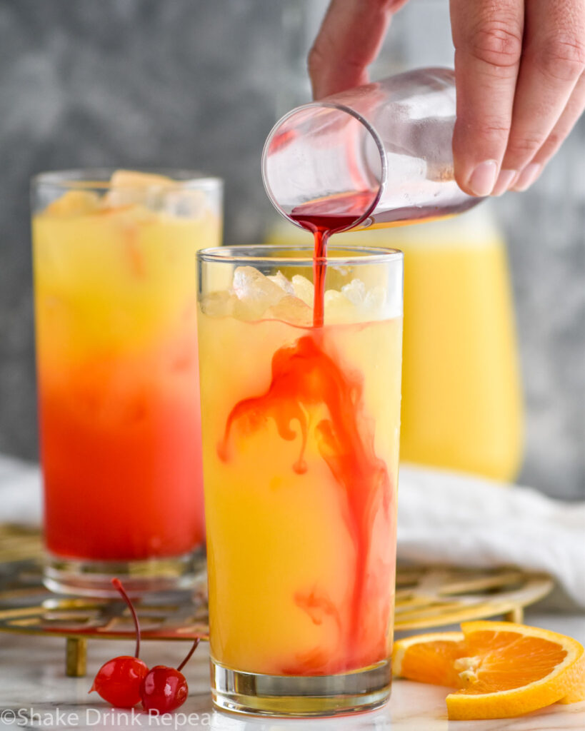 man pouring grenadine into a glass of tequila sunrise and ice surrounded by orange slices, cherries, and a pitcher of orange juice