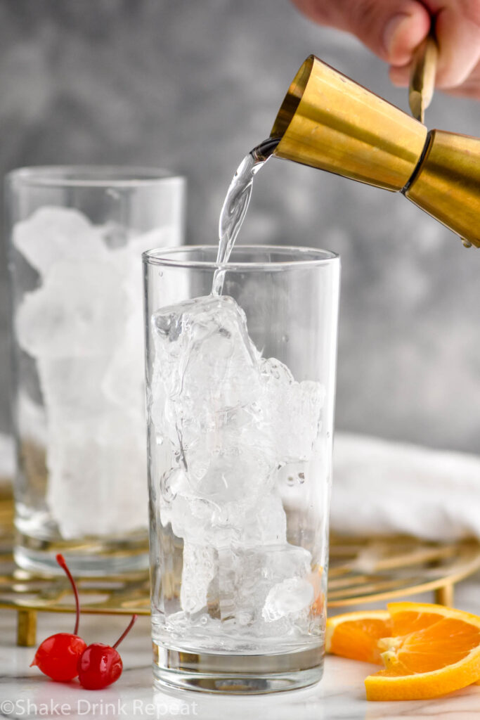 jigger of tequila pouring into a glass of ice to make a tequila sunrise surrounded by orange slices and cherries for garnish