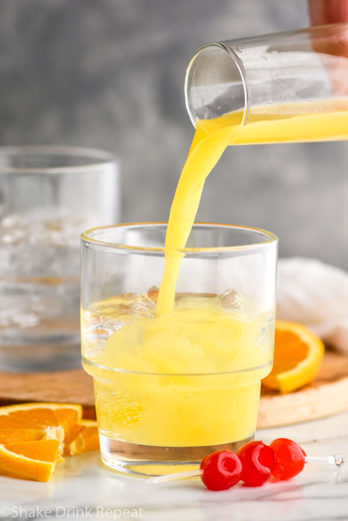 pouring orange juice into a glass of tequila sunset recipe surrounded by cherries and orange slices