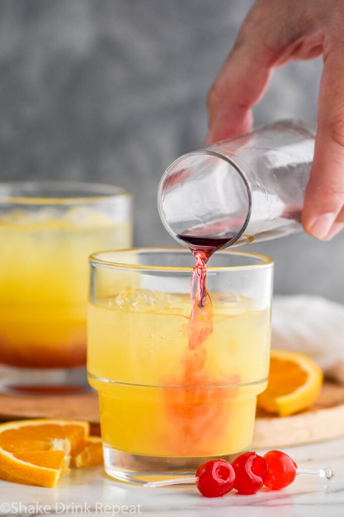 man's hand pouring blackberry brandy into a glass of tequila sunset ingredients and ice surrounded by orange slices and cherries