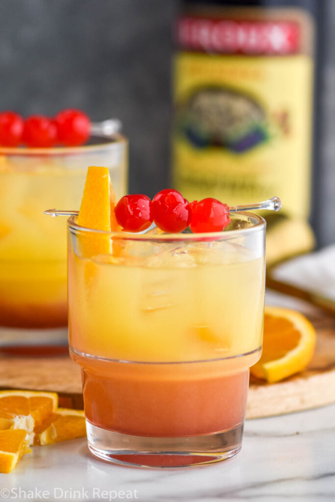 two glasses of tequila sunset with orange and cherry garnish and bottle of blackberry brandy in the background