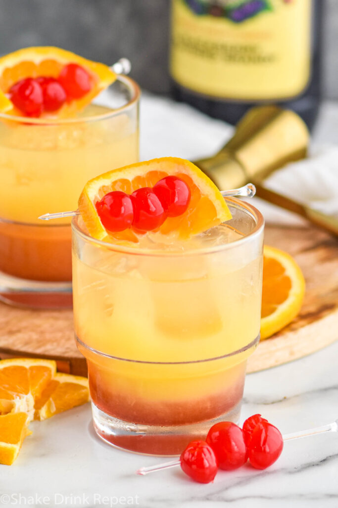 two glasses of tequila sunset with ice and cherries and orange slice garnish with bottle of blackberry brandy in the background