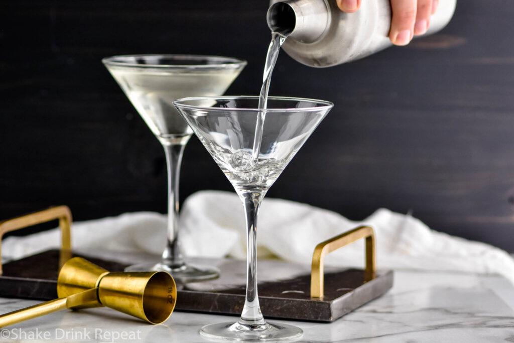 man pouring shaker of Vesper Martini ingredients into a martini glass surrounded by jigger and glass of vesper martini