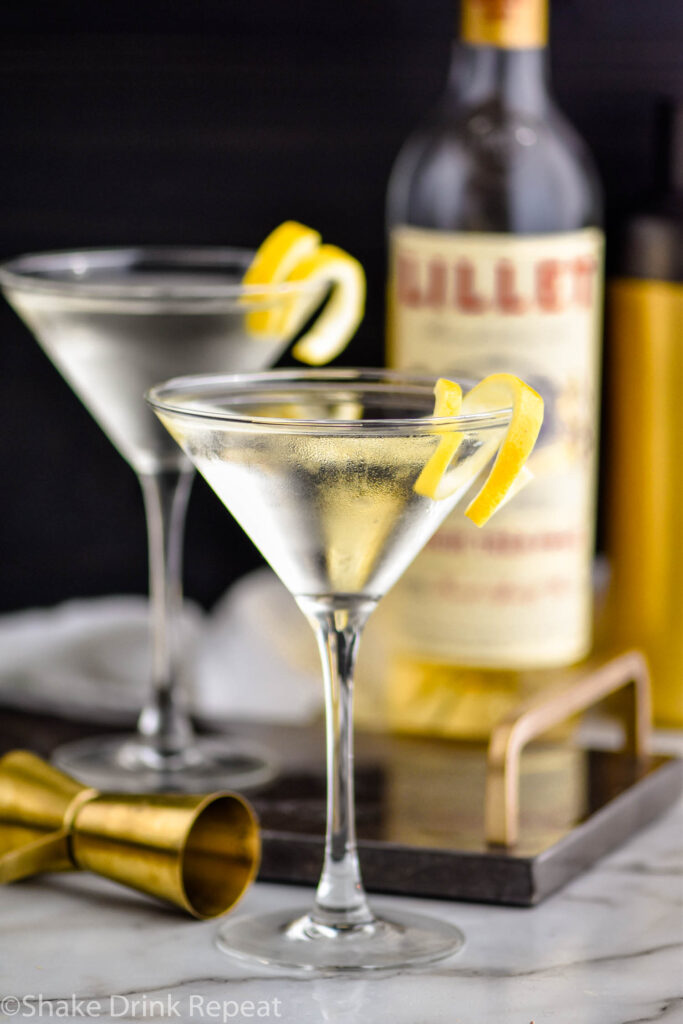 two martini glasses of Vesper Martini with lemon twist garnish surrounded by bottle of Lillet Blanc and jigger