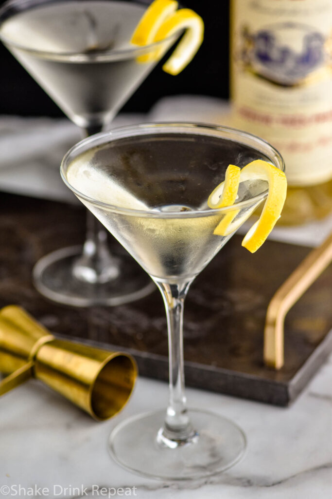 two martini glasses of Vesper Martini with lemon twist garnish surrounded by jigger and bottle of Lillet Blanc