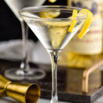 two martini glasses of Vesper Martini with lemon twist garnish and bottle of Lillet Blanc and jigger in the background