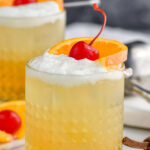 two glasses of Whiskey Sour with ice, frothy egg white, and cherry and orange slice garnish