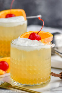 two glasses of Whiskey Sour with ice, frothy egg white, and cherry and orange slice garnish