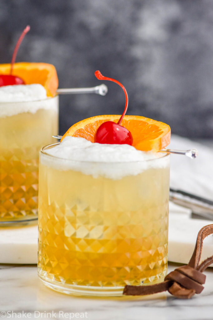 two glasses of Whiskey Sour with ice, frothy egg white, and cherry and orange slice garnish