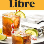 two glasses of Cuba Libre with ice, slices of lime, and straws surrounded by a jigger and lime wedges