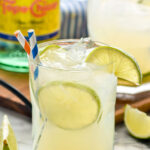 Glass of Ranch Water cocktail with ice, straws, and slices of lime with second glass of Ranch Water cocktail and bottle of Topo Chico in the background