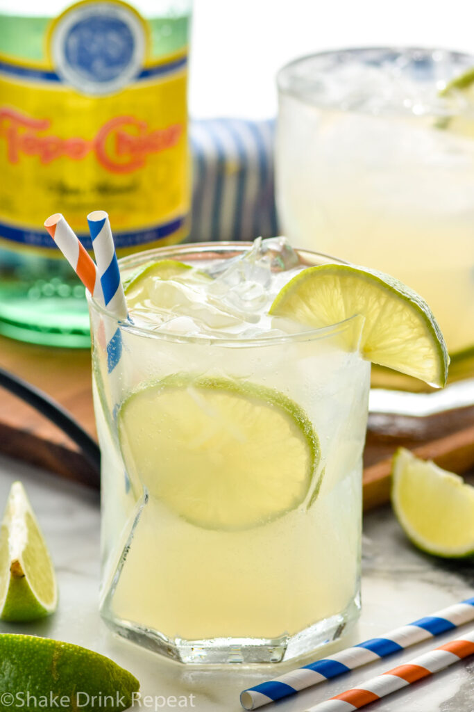 Glass of Ranch Water cocktail with ice, straws, and slices of lime with second glass of Ranch Water cocktail and bottle of Topo Chico in the background