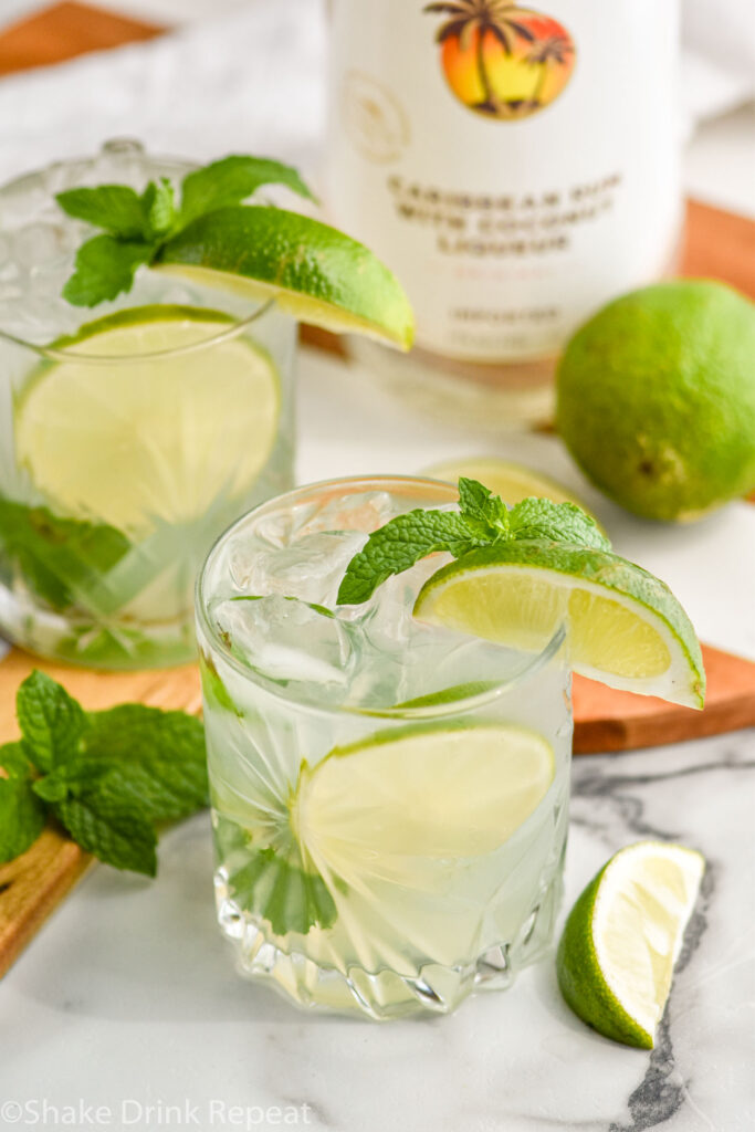 two glasses of Cojito with ice, mint leaves, and slices of lime with bottle of Malibu rum in the background