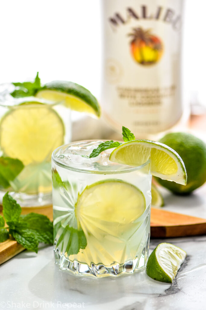 glass of cojito with ice, mint leaves, and lime slices with bottle of Malibu rum in the background