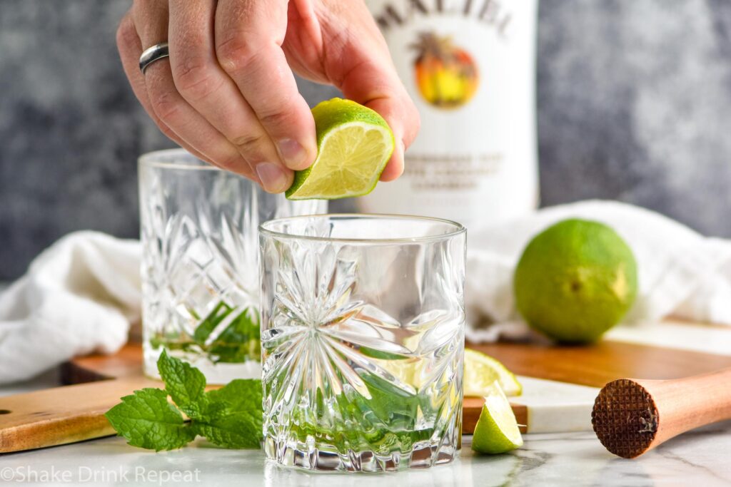 man's hand squeezing juice from a lime into a glass to make a Cojito recipe surrounded by mint leaves and bottle of Malibu rum.