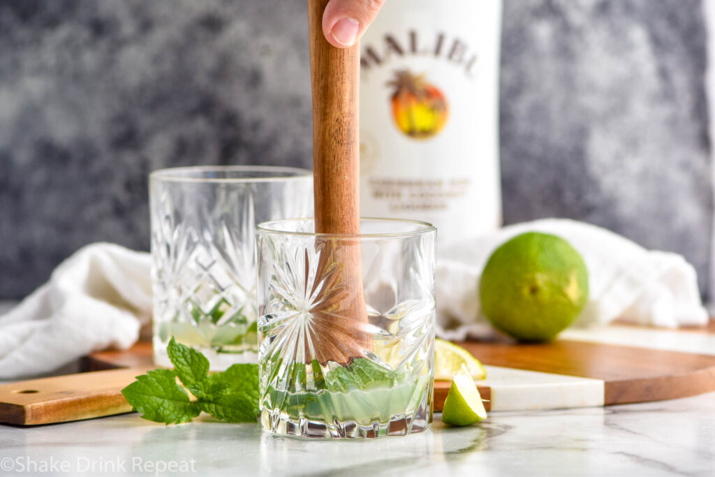 man's hand muddling mint leaves in a glass to make a Cojito recipe surrounded by mint leaves, slices of lime, and bottle of Malibu rum