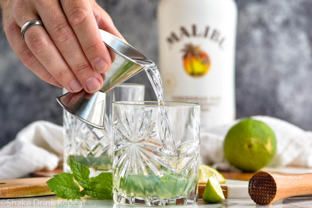 man's hand pouring jigger of coconut rum into a glass of Cojito recipe ingredients surrounded by fresh mint leaves, slices of lime, muddler, and bottle of Malibu rum.
