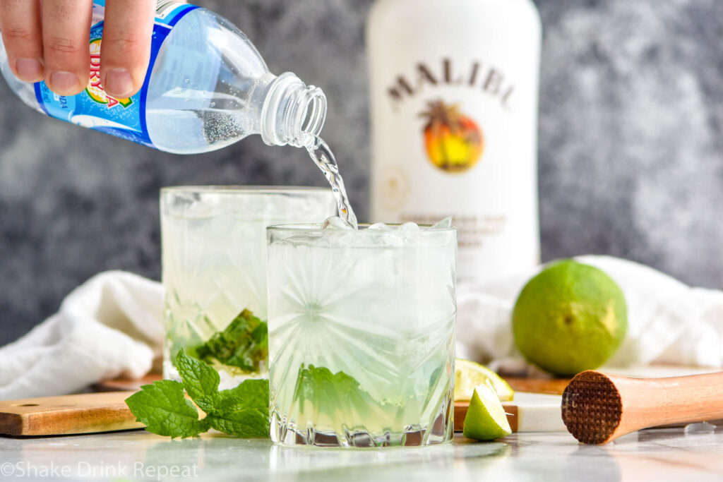 man's hand pouring bottle of club soda into a glass of Cojito recipe surrounded by mint leaves and slices of lime with bottle of Malibu rum in the background